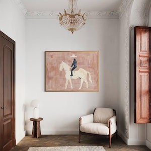 May include: An abstract painting of a person riding a white horse, set against a pink background. The artwork is framed in gold and hangs on a white wall. The room features a chandelier, a side table, and a cream-colored armchair.