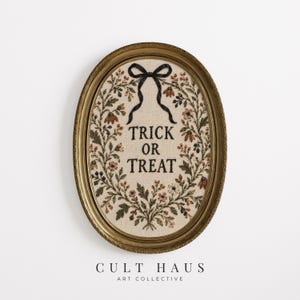 May include: An oval gold frame containing an embroidered piece on cream fabric. The embroidery features a black bow at the top, a floral wreath in shades of red, orange, and green, and the black text TRICK OR TREAT centered in the middle.