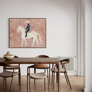 May include: A framed abstract painting depicts a rider on a white horse against a muted pink background. The rider is wearing a dark outfit and a white hat. The artwork is displayed in a room with a wooden table and chairs.