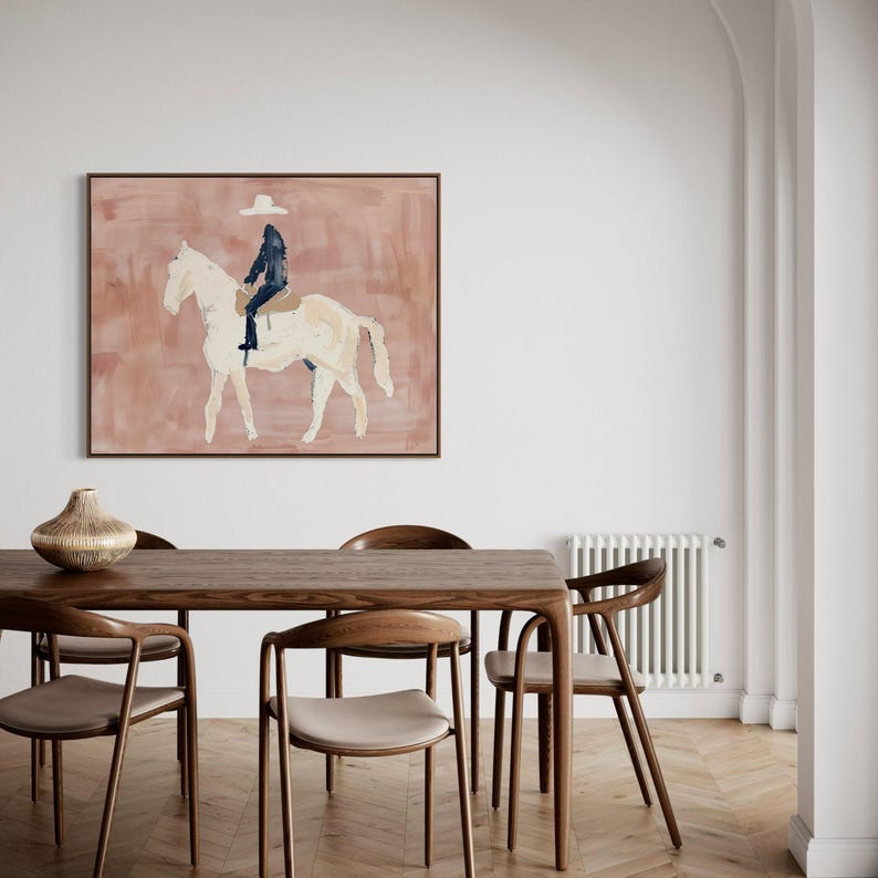 May include: A framed abstract painting depicts a rider on a white horse against a muted pink background. The rider is wearing a dark outfit and a white hat. The artwork is displayed in a room with a wooden table and chairs.