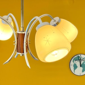 Könnte beinhalten: Ein Kronleuchter im Vintage-Stil mit fünf Lichtern. Die Leuchte hat eine Messingoberfläche und einen Holzakzent. Die Lampenschirme sind cremefarben mit einem Sternmuster. Im Hintergrund befindet sich eine Uhr mit einer Weltkarte.