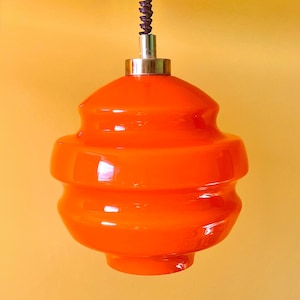 Könnte beinhalten: Eine leuchtend orangefarbene Glas-Hängeleuchte mit abgestufter, abgerundeter Form. Die Leuchte hat eine glatte, glänzende Oberfläche und ist an einem dunklen, spiralförmigen Kabel befestigt, das an einer silberfarbenen Metallkappe oben am Glasschirm sitzt.