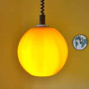 Può includere: Una lampada a sospensione sferica in vetro giallo brillante è appesa a un cavo nero. Un piccolo orologio rotondo con un disegno della mappa del mondo è montato sulla parete gialla. L'orologio ha le lancette nere.