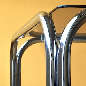 May include: Close-up of a chrome metal bar cart with a dark glass top. The cart has a curved, arched design with a reflective surface. The background is a solid, warm yellow colour.