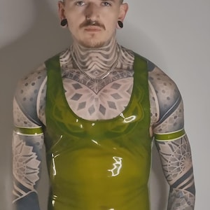 May include: An olive-green, shiny tank top with a scoop neck and armholes. The top has a glossy finish and is worn with black trousers. The person wearing the top has intricate tattoos on their arms, chest, and neck.