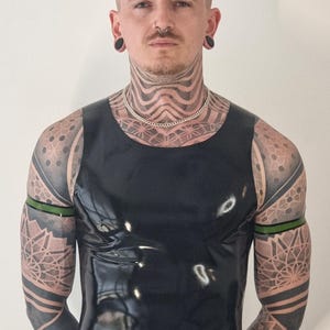 May include: A black, shiny sleeveless top. The person in the image has pink hair, ear gauges, and detailed tattoos on the neck, shoulders, and arms. A silver chain necklace is also visible.