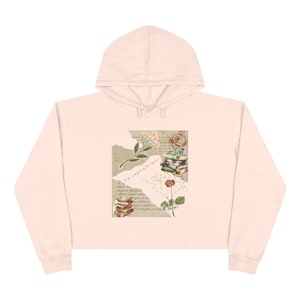 May include: A light peach-colored cropped hoodie featuring a central graphic collage. The design includes stacks of books, botanical illustrations of flowers, and torn paper textures with the printed Spanish text Un capítulo más.