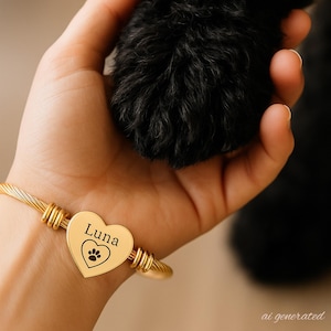 May include: A gold-toned cable wire bracelet featuring a central heart-shaped charm. The charm is engraved with the name Luna above a small paw print icon inside a heart outline. The bracelet is worn on a wrist near a black furry animal paw.