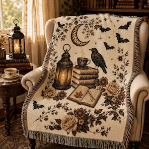 Puede incluir: Una manta tejida con fondo crema y un patrón marrón oscuro de una luna creciente, murciélagos volando, un cuervo negro sobre una pila de libros, una linterna encendida, un libro abierto con una hoja y detalles de rosas con flecos.