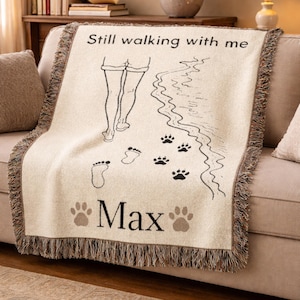 May include: Cream-colored woven throw blanket with a fringed edge. The blanket features a black and white illustration of a person walking on a beach, with paw prints and the text "Still walking with me" and "Max".