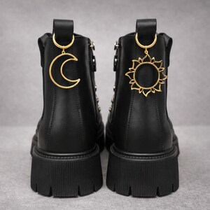 Könnte beinhalten: Schwarze Stiefeletten mit dicken Sohlen. Jede Stiefel hat einen goldenen Anhänger, einer in Form eines Halbmonds und der andere in Form einer Sonne. Die Stiefel haben einen Reißverschluss und stehen vor grauem Hintergrund.