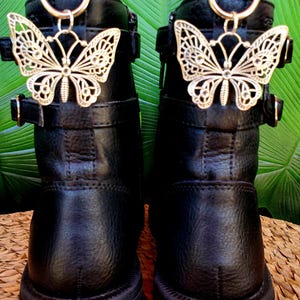 Peut inclure: Paire de bottines noires avec des breloques papillon dorées. Les bottines ont une semelle épaisse, des boucles et une fermeture éclair. Les breloques papillon sont fixées en haut des bottines avec un anneau doré.