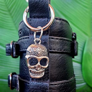 Op de afbeelding: Zwart leren accessoire met zilverkleurige hardware, met een schedelbedel. Het accessoire heeft een riem met gespen en een metalen ring. De schedelbedel is gedetailleerd met getextureerde oppervlakken en open oogkassen.