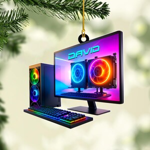 May include: A computer-themed ornament featuring a desktop computer, keyboard, and tower with colorful LED lights. The monitor displays the name "DAVID" in blue. The ornament hangs from a gold string and black ring, with a green background.