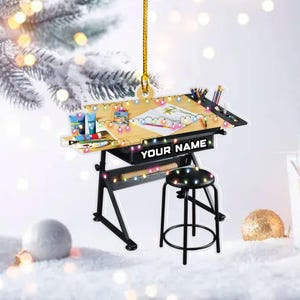 May include: A Christmas ornament shaped like a drafting table with a stool. The table is decorated with colorful lights and art supplies. The text "YOUR NAME" is displayed on the front of the table.