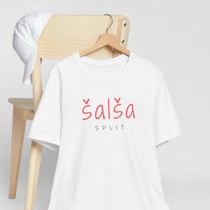 May include: A white t-shirt with the word "šalša" in red script above the word "SPLIT". A white baseball cap rests on a light wooden chair. The t-shirt hangs on a wooden hanger.