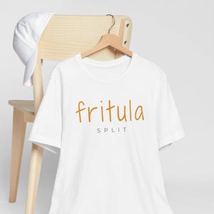 May include: A white t-shirt with the word "fritula" in a handwritten style, and the word "split" below it, in a brown color. The t-shirt is hanging on a wooden hanger. A white baseball cap rests on the back of a wooden chair.