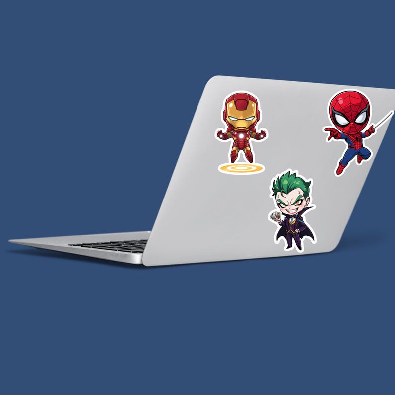 May include: A silver laptop with four cartoon stickers: Iron Man in red and gold, Spider-Man in red and blue, and the Joker in purple and green. The stickers are placed on the laptop's lid.