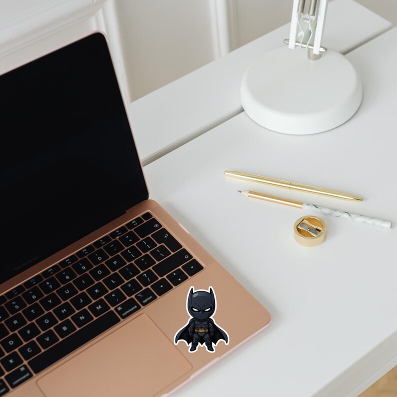 May include: A sticker featuring a chibi-style Batman character in a black suit with a yellow utility belt. The sticker is placed on a rose gold laptop, with a white desk, pens, and a pencil sharpener in the background.