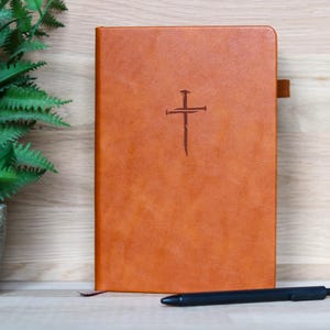 May include: A brown leather journal with a cross symbol embossed on the cover. A black pen rests on the wooden surface in front of the journal. A small green plant in a gray pot is to the left.