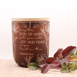 May include: A brown glass candle jar with a wooden lid, featuring white floral designs and the text "may the GOD OF HOPE fill you with JOY AND PEACE as you trust in Him ROMANS 15:13". Decorative leaves are placed near the candle.
