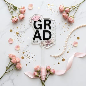 Peut inclure: Un cake topper de graduation avec le mot "GRAD" en noir, l'année "2026" et une illustration de diplôme. Le topper est décoré d'une toque rose et du nom "ALEXA". L'image est entourée de roses roses, de confettis dorés et d'un collier de perles.