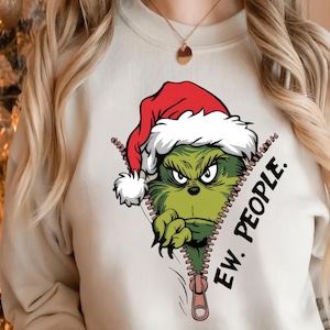 May include: A beige sweatshirt featuring a green Grinch character wearing a Santa hat. The Grinch is peeking out from a zipper design, with the words "EW. PEOPLE." printed on the right side of the zipper.