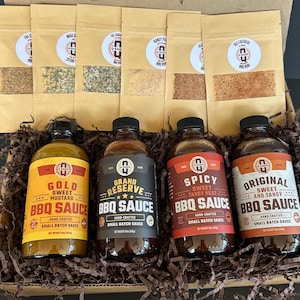 Könnte beinhalten: Ein BBQ-Geschenkset mit vier Saucenflaschen und fünf Gewürzsäckchen. Die Saucen sind mit "Gold Sweet Mustard", "Grand Reserve", "Spicy Sweet Tangy Heat" und "Original Sweet and Tangy" beschriftet. Die Gewürze sind in braunen Papiertüten.