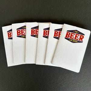 May include: Several white notebooks are displayed, each featuring the text "THE BEER LOG BOOK" in red and navy blue. The notebooks are arranged on a dark surface, highlighting the cover design.