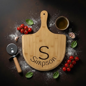 Può includere: Una tavola di legno per pizza con manico, incisa con la lettera "S" e il nome "Simpson". La tavola è circondata da ingredienti come pomodori, basilico, aglio e un tagliapizza, suggerendo un tema culinario.
