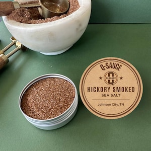 May include: A small, open, silver tin filled with brown seasoning, next to a round label that reads "Q-SAUCE Hickory Smoked Sea Salt." A white marble mortar and pestle with a scoop and more seasoning is in the background. The background is green.