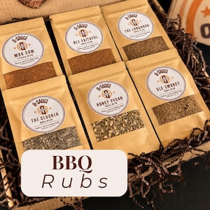 Op de afbeelding: Een verzameling van zes kleine batches BBQ-rub-pakketten in een geschenkdoos. De pakketten zijn bruin met ronde etiketten en namen als "Moo Cow" en "Honey Pecan". De tekst "BBQ Rubs" wordt weergegeven op een witte rechthoek.