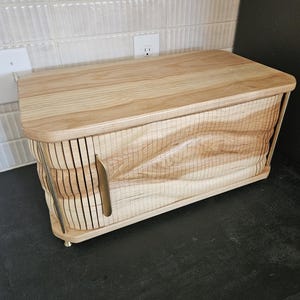 May include: A light-colored wooden bread box with a curved top and a unique, textured front design. The box features a handle and rests on small, gold-colored feet. The wood grain is visible throughout the design, adding to its natural aesthetic.
