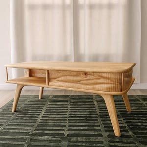 May include: A light-colored wooden coffee table with a lower shelf and curved legs. The table has a natural wood grain finish and a rectangular shape. It sits on a dark green rug with a geometric pattern. The background includes a white curtain.