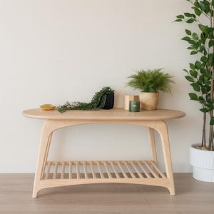 May include: A light-colored wooden coffee table with an oval top and a slatted shelf. The table has curved legs and is decorated with small potted plants and a decorative item. A tall plant is in the background.