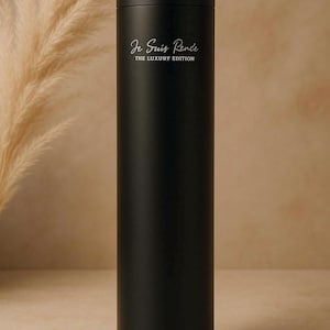 Puede incluir: Un difusor cilíndrico negro con base redonda. El texto "Je Suis Rende THE LUXURY EDITION" está impreso en blanco en la parte delantera. Una pequeña luz azul es visible en la base. El fondo es beige neutro.