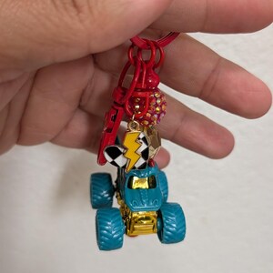 May include: A red keychain with a blue and gold monster truck charm. The keychain also features a lightning bolt charm, a checkered flag charm, and a red charm with a small red bead. The monster truck has large blue tires.