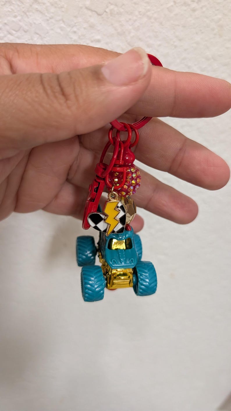 May include: A red keychain with a blue and gold monster truck charm. The keychain also features a lightning bolt charm, a checkered flag charm, and a red charm with a small red bead. The monster truck has large blue tires.