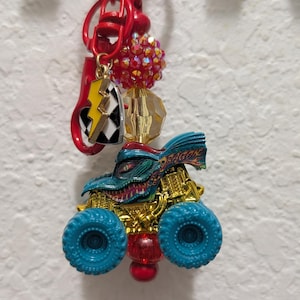 May include: A red keychain with a monster truck charm, a lightning bolt charm, and decorative beads. The monster truck is blue and gold with blue wheels. The keychain is hanging on a black hook.