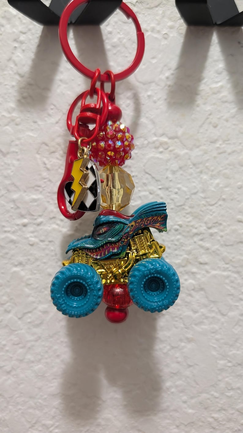 May include: A red keychain with a monster truck charm, a lightning bolt charm, and decorative beads. The monster truck is blue and gold with blue wheels. The keychain is hanging on a black hook.
