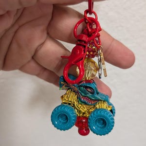 May include: A red keychain with a monster truck charm. The truck has teal tires, a gold body, and a teal and gold monster face. The keychain has a red clasp and a cluster of small charms.