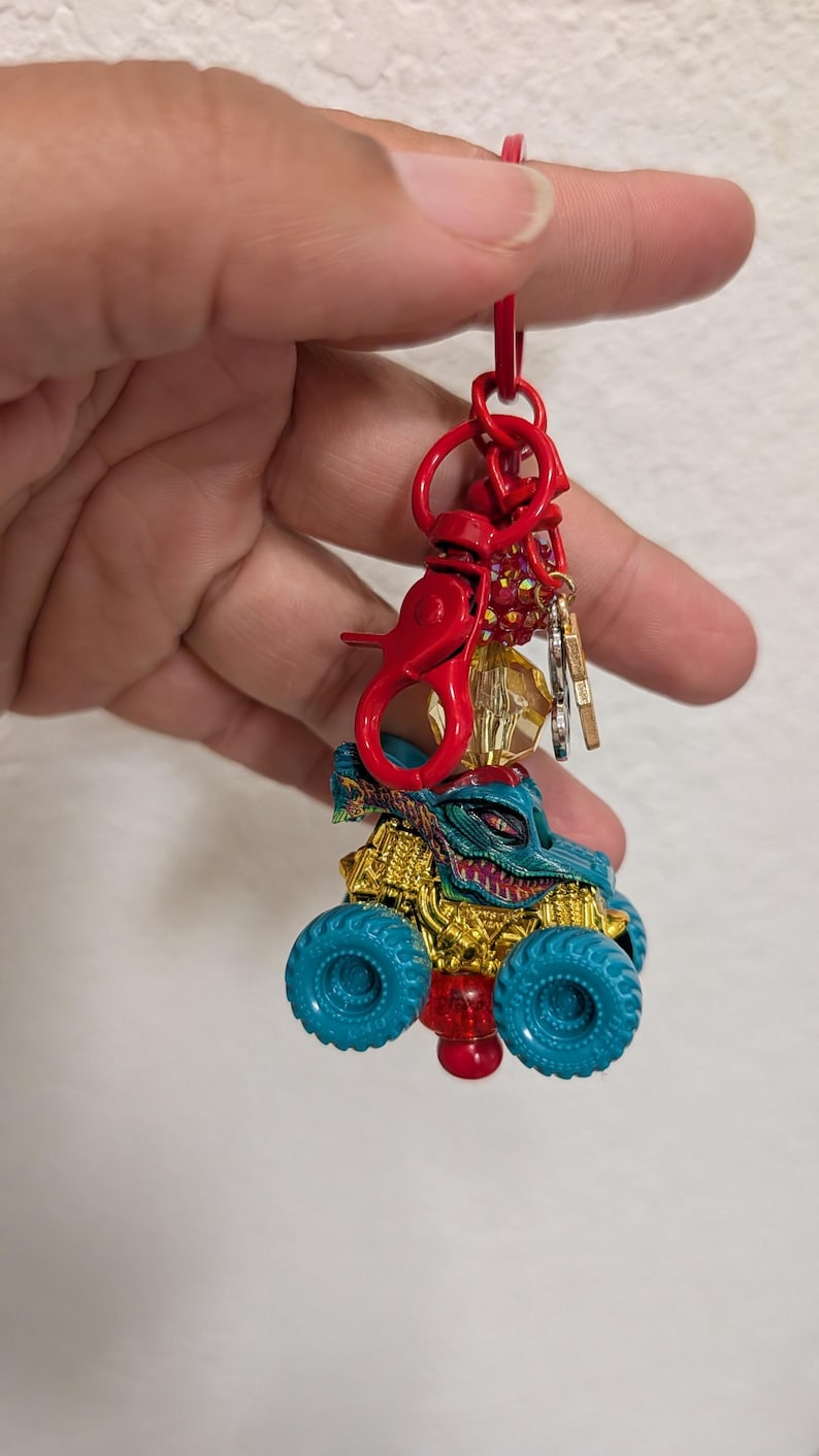 May include: A red keychain with a monster truck charm. The truck has teal tires, a gold body, and a teal and gold monster face. The keychain has a red clasp and a cluster of small charms.