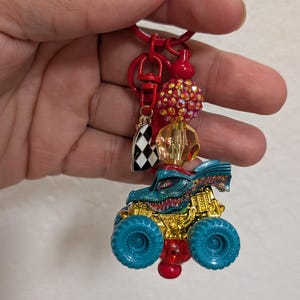 May include: A red keychain with a monster truck charm. The truck is teal and gold with teal wheels. The keychain also features a checkered flag charm, red beads, and a large, sparkly red bead.