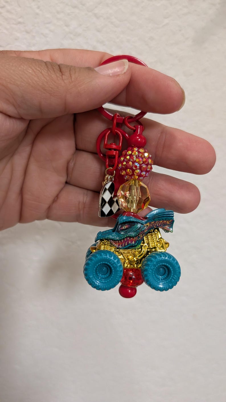 May include: A red keychain with a monster truck charm. The truck is teal and gold with teal wheels. The keychain also features a checkered flag charm, red beads, and a large, sparkly red bead.