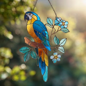 Puede incluir: Vidriera de loro con colores azul, amarillo y naranja. El loro está posado en una rama marrón con flores azules y hojas verdes. Incluye una cadena decorativa para colgar.
