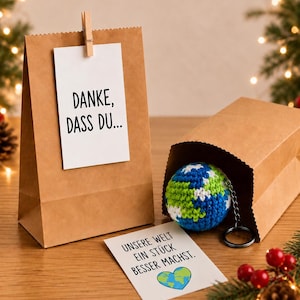 Könnte beinhalten: Eine braune Papiertüte mit einem angeklammerten weißen Schild mit der Aufschrift "DANKE, DASS DU..." und eine zweite Tüte mit einem gehäkelten Globus-Schlüsselanhänger. Vorne liegt eine Karte mit "UNSERE WELT EIN STÜCK BESSER MACHST." und einem Erdenherz.