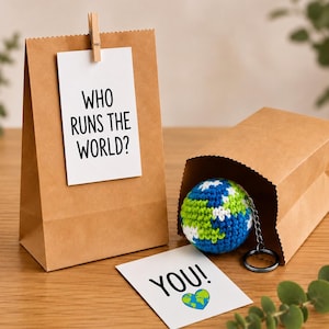Könnte beinhalten: Eine braune Papiertüte mit einer angeklammerten weißen Karte mit der Aufschrift WHO RUNS THE WORLD? Eine zweite Tüte enthält einen kugelförmigen, gehäkelten Globus-Schlüsselanhänger in Blau, Grün und Weiß. Vorne liegt eine Karte mit YOU! und Herz-Globus.