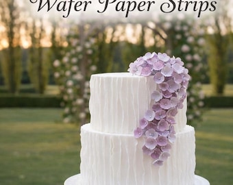 May include: A two-tier white cake with a vertical ruffled texture, decorated with a cascading cluster of light purple wafer paper flowers. The text "Wafer Paper Strips" appears at the top, and "Make your very own professional cake!" at the bottom.