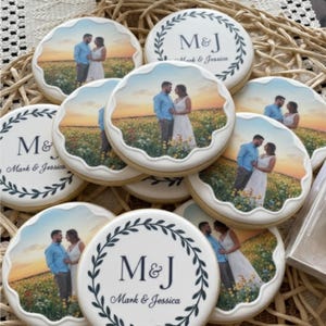 May include: A collection of decorated sugar cookies. Some cookies feature a couple in a field at sunset, while others display the initials "M&J" with "Mark & Jessica" below, surrounded by a wreath design. The cookies have a white scalloped edge.