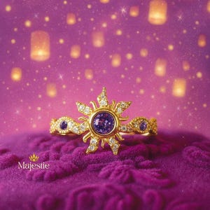 Peut inclure: Une bague en or avec un motif de soleil, orn&eacute;e d'une pierre pr&eacute;cieuse violette centrale et de diamants. La bague est pos&eacute;e sur une surface de velours violet, avec un fond de lanternes flottantes et de lumi&egrave;res scintillantes. Le mot "Majestie" est en bas.